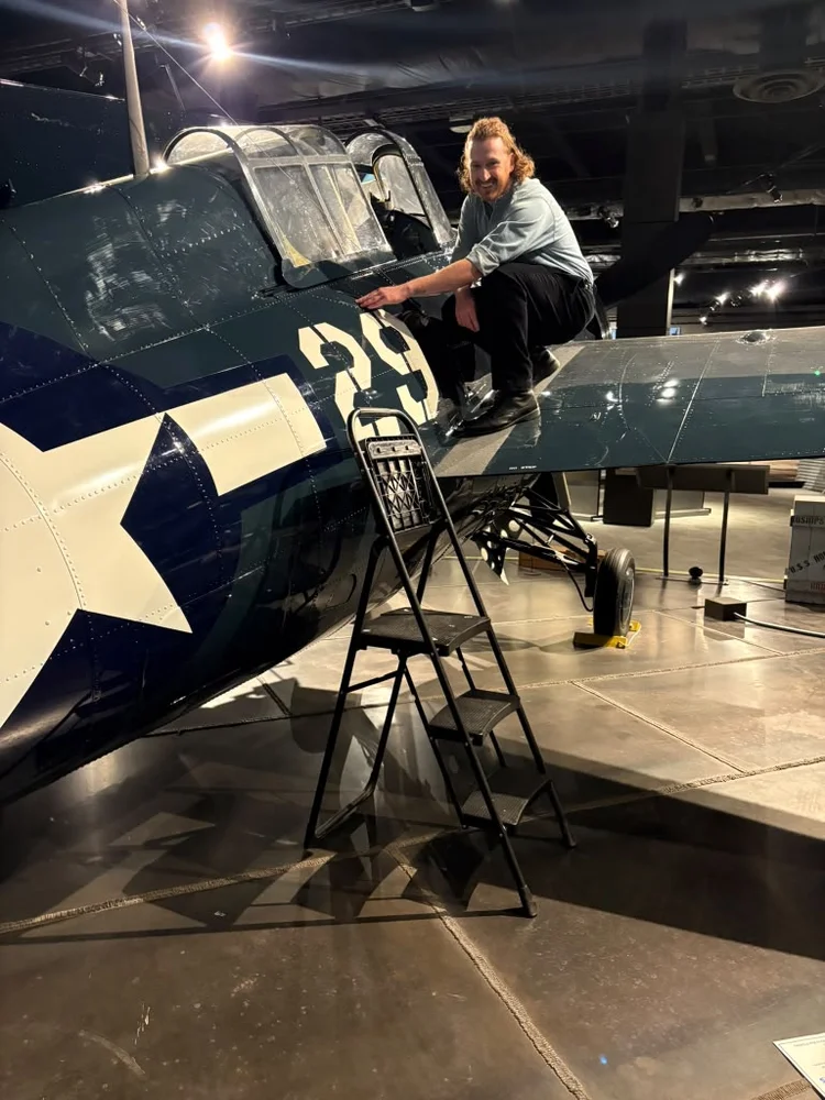 Evan Weber crouching on the wing of a WWII Navy aircraft in a museum hangar